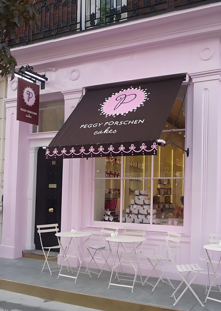 The iconic pastel pink facade of Peggy Porschen Cakes in London's Notting Hill, with a branded black awning and outdoor seating.