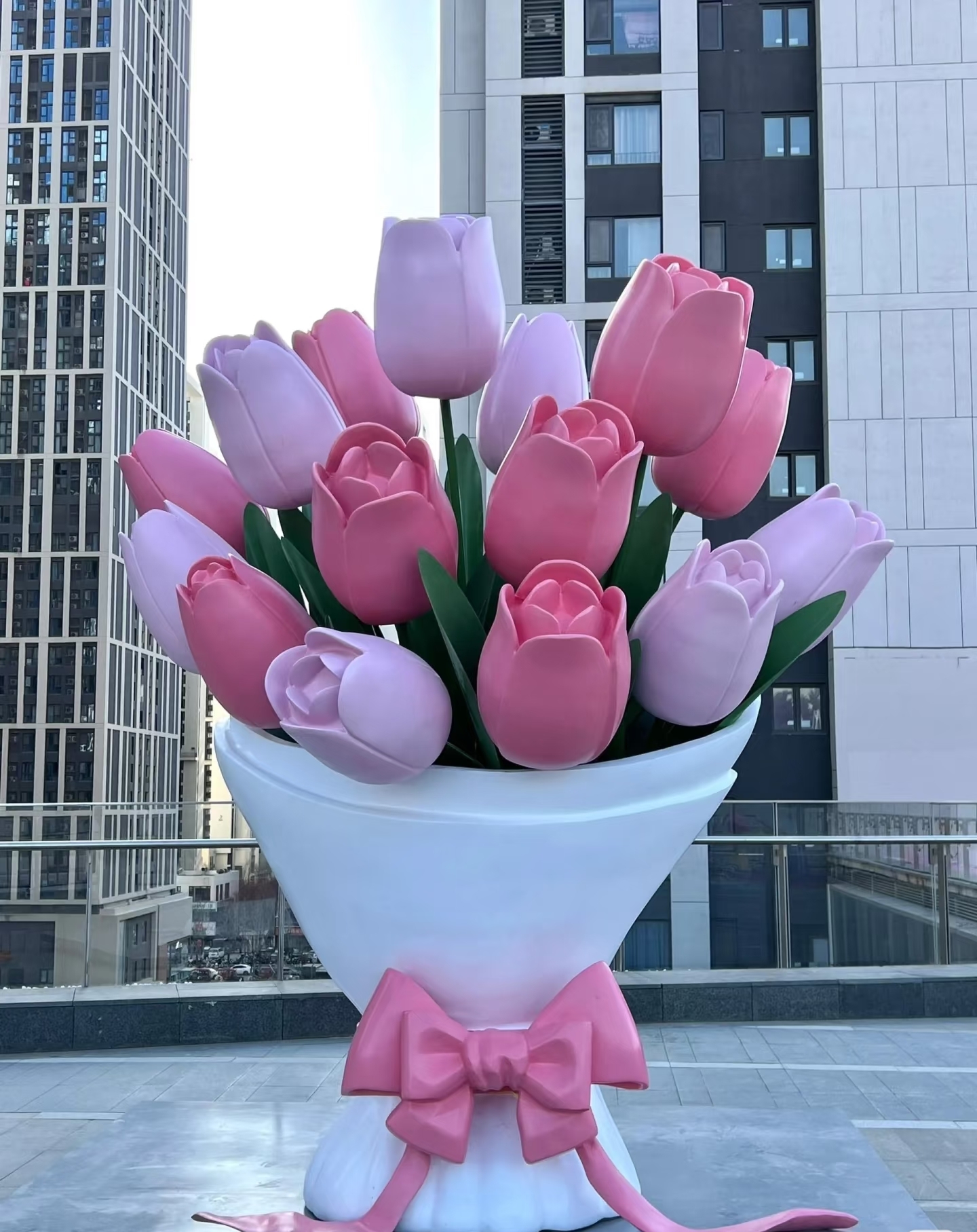 Giant tulip hand‑held fiberglass bouquet sculpture in pink‑purple tones on an outdoor urban terrace with high‑rise buildings
